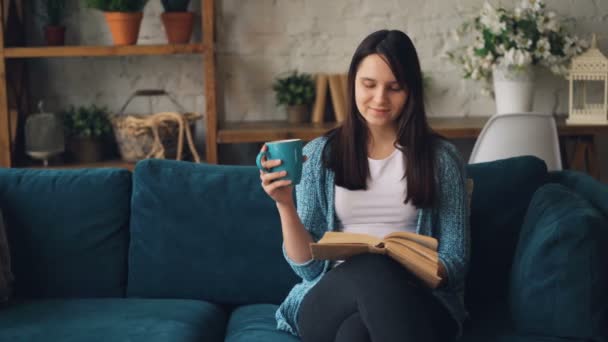 Menina bonita está lendo livro sentado no sofá em casa com xícara de chá e relaxar desfrutando de solidão e paz. Educação, geração Y e conceito de casa . — Vídeo de Stock