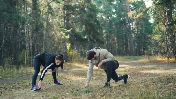 Mladý muž a žena jsou školení v parku společně natahovat nohy teplý podzimní den nosit módní sportovní oblečení. Lidé, sport a volný čas koncepce. — Stock video