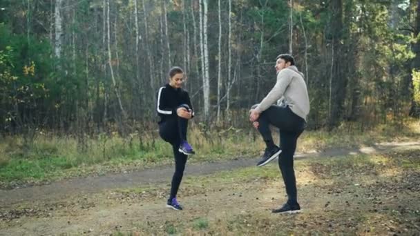 Barbudo joven está haciendo deportes en el parque con su novia estirando las piernas haciendo calentamiento sonriendo y hablando. Hermosa naturaleza de otoño está en el fondo . — Vídeo de stock