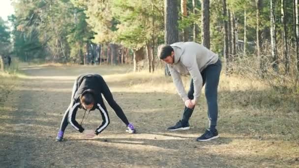 Un par de amigos macho y hembra están haciendo deportes al aire libre inclinándose hacia adelante durante el calentamiento en el día soleado de otoño. Vida sana, gente y concepto de la naturaleza . — Vídeos de Stock