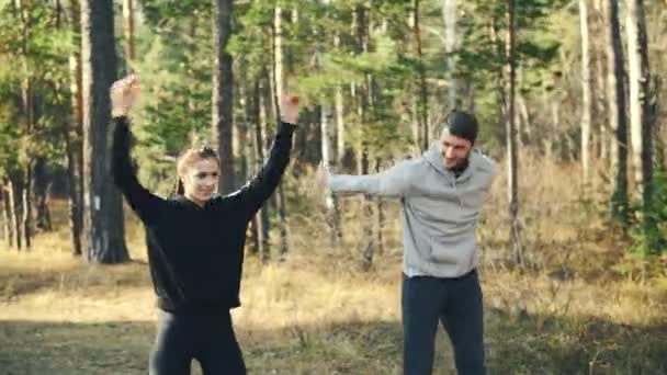 Attractive young woman in sportswear is exercising in park with her boyfriend doing warm-up rotating arms talking and smiling. Healthy lifestyle and nature concept. — Stock Video