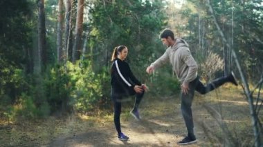 Kız ve adam birlikte hareket konuşuyor ve ağaçları arasında yol üzerinde duran gülümseyen ısınma-up eklem bacağı orman spor yapıyoruz. İnsanlar spor giyim üzerinde var.