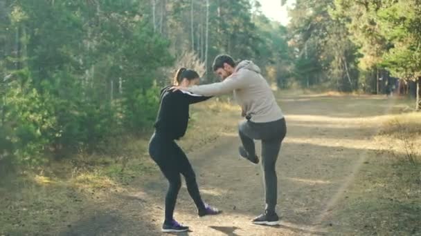 Man and woman are enjoying sports in park holding each others shoulders and moving legs warming-up joints standing on path with green trees around them. — Stock Video