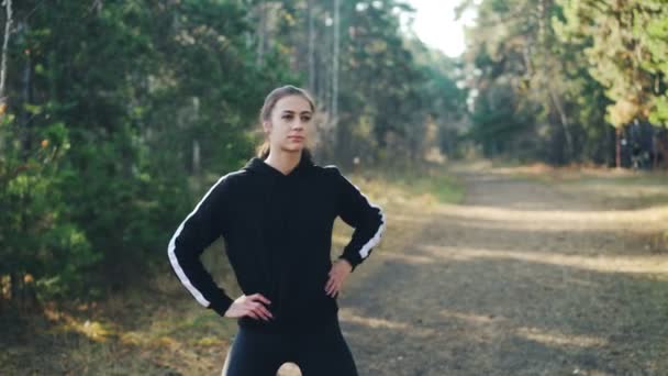 Bella bruna in abbigliamento sportivo moderno si allena da solo nel parco piegando e muovendo braccia e corpo godendo di aria fresca, attività fisica e natura . — Video Stock