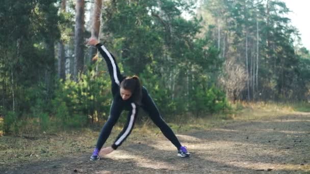 Attractive young sportswoman is doing sports in park on sunny autumn day wearing modern black tracksuit. Slim girl is bending forward and moving arms. — Stock Video