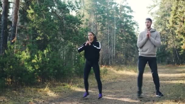 Alegre estudiantes están haciendo deportes juntos en el parque en cuclillas y sonriendo disfrutando de los ejercicios y la naturaleza. Hombre y mujer están usando chándales modernos . — Vídeo de stock