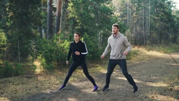 Los jóvenes sonrientes mujer y hombre están saltando en el bosque haciendo deportes con chándales y zapatillas de deporte que se divierten y cuidan de la salud. Concepto naturaleza y ejercicios . — Vídeos de Stock