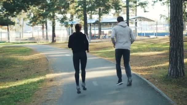 Rear view of beautiful couple man and woman running in park together wearing trendy sportswear and sneakers. Active youth, healthy people and recreation concept. — Stock videók