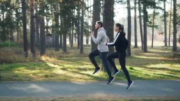 Sportovní mladá žena vykonává venku s kamarádkou mužského běhu a skákání v parku mezi zelenými stromy. Dětí a mládeže, šťastní lidé a příroda koncepce. — Stock video