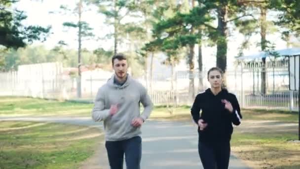 Dolly shot of male and female students running in park together on autumn day wearing modern sportswear. Evergreen pine trees and grass are visible. — Stock Video