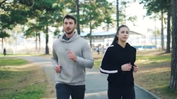 Bearded young man is jogging with his female friend in park exercising outdoors wearing modern sportswear concentrated on running. Healthcare and people concept. — Stock Video