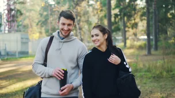 Fiatal és vonzó lány beszél, hogy a barátja, és nevetve, séta a parkban, a sport-táskák szabadtéri edzés után. Beszélgetés, szórakoztató és aktív ifjúsági koncepciót. — Stock videók