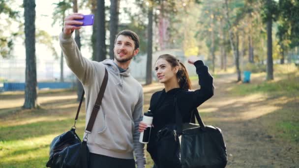 Счастливые друзья спортивные молодые люди делают селфи с помощью смартфона после тренировки в парке. Мужчина и женщина держат сумки и позируют перед камерой . — стоковое видео
