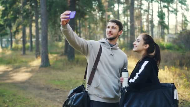 Молоді люди чоловік і жінка беруть селфі в парку з камерою смартфона, позуючи з пляшкою води і спортивними сумками. Дівчина сміється і показує біцепс . — стокове відео