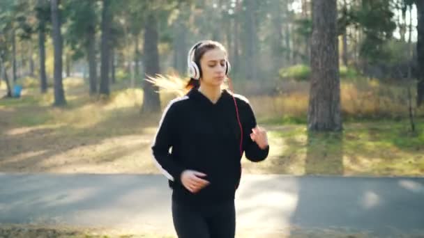 Dolly shot di carina bruna che fa jogging nel parco e ascolta la radio in cuffia che si esercita da sola. Moderna tecnologia, assistenza sanitaria e concetto di ricreazione . — Video Stock