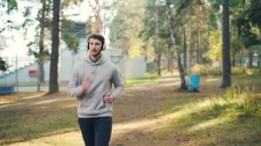 Kulaklık ile müzik dinlemek ve bazen kameraya bakarak park, koşu öğrenci portresi. Adam son moda spor giyim giymek ve modern cihaz kullanarak.
