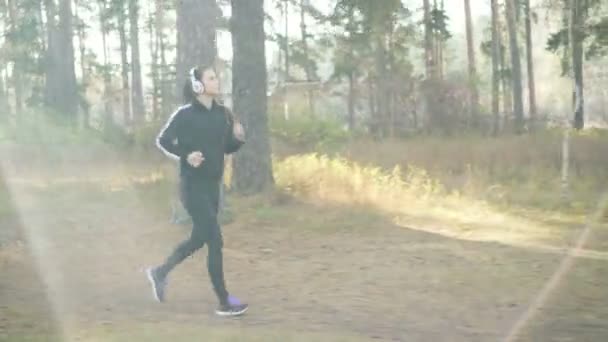 Mooi meisje is joggen in het park en het luisteren naar muziek via de koptelefoon op zonnige herfstdag dragen zwart trainingspak moderne sportkleding. Jeugd en lichaamsbeweging concept. — Stockvideo