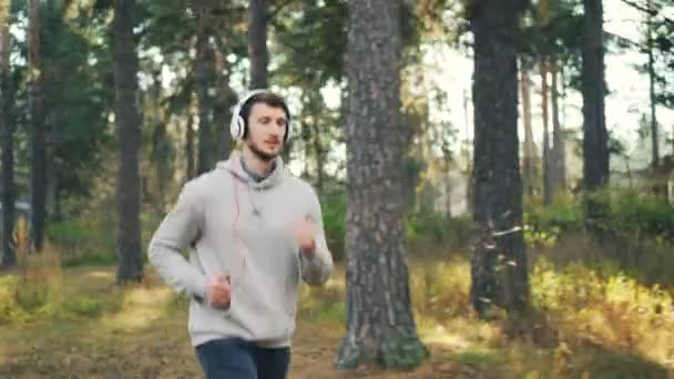 Dolly scatto di bel giovane uomo che fa jogging nel parco e ascolta musica attraverso le cuffie godendo di canto e attività fisica. Cultura giovanile e concetto di sport . — Video Stock