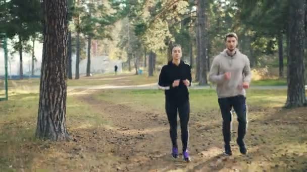 Dolly shot of attractive girl and guy jogging in park together focused on practice. Femme porte un sweat-shirt et des leggings, homme porte un survêtement . — Video