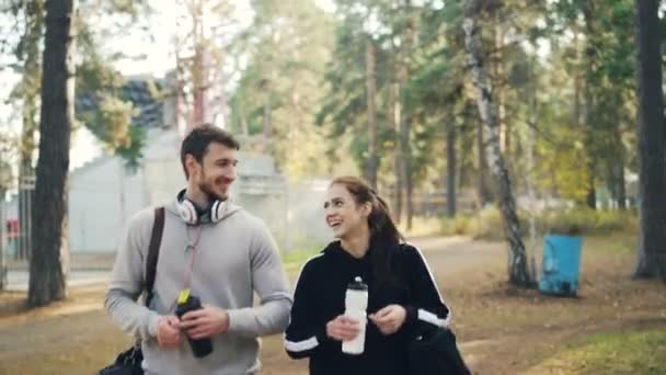 Il giovane bell'uomo in cuffia sta parlando con la sua amica e ride camminando nel parco con borse sportive dopo l'allenamento all'aperto poi facendo il cinque . — Video Stock