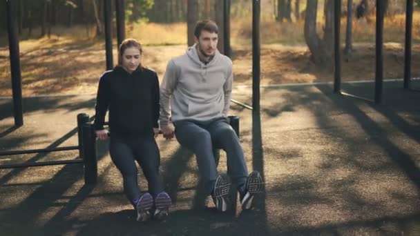 Die junge Frau trainiert mit ihrem sportlichen Freund draußen auf dem Sportplatz mit den Armen am Stufenbarren. Paar, Sport und Naturkonzept. — Stockvideo