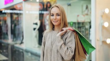 Kameraya bakarak ve ayakta alışveriş merkezinde kağıt torba tutarak yalnız gülümseyen trendy giyim oldukça uzun saçlı sarışın portresi. İnsanlar ve dükkanlar kavramı.