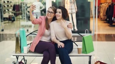 Genç ve güzel kadınlar alışveriş merkezi bankta oturmuş ve having fun smartphone kamera kullanarak selfie alıyor. Modern teknoloji, dükkanlar ve arkadaşlar kavramı.