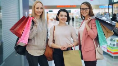Trendy giyim ve aksesuarları büyük kameraya bakarak ve gülümseyen alışveriş merkezinde alışveriş torbaları tutmak yakışıklı kızlar portresi. İnsanlar ve mağaza konsepti.