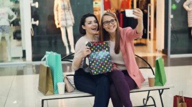 Şirin kızlar selfie alışveriş merkezi kağıt poşet ve butik bankta oturmuş son moda el hareketleri ile poz alarak. Kadın eğlenmek ve gülmek.
