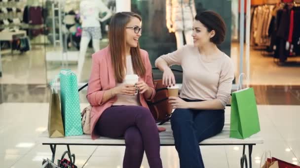 Chicas felices están socializando charlando y riendo sentado en el banco en el centro comercial con bebidas para llevar y bolsas de compras. Los escaparates con prendas son visibles . — Vídeo de stock