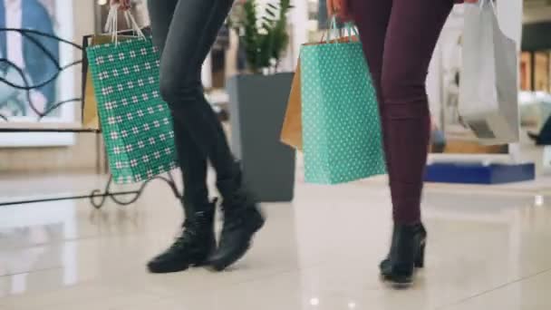 Amigas estudantes estão andando em shopping segurando sacos de papel em mãos desfrutando venda temporada. Pernas femininas, loja de luz interior e roupas são visíveis . — Vídeo de Stock