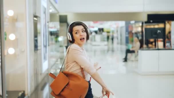 Feliz hembra adicta a las compras se divierte en el centro comercial escuchando música en los auriculares, bailando con bolsas brillantes y riendo apuntando a los productos en los escaparates . — Vídeo de stock