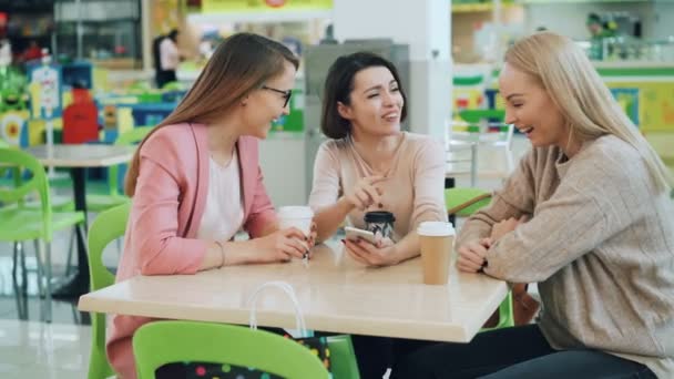 Amigos felices mujeres guapas están mirando a la pantalla del teléfono inteligente y hablando sentado en la cafetería a continuación, haciendo chocar los cinco y riendo. Joven moderna, dispositivos y concepto de amistad . — Vídeo de stock