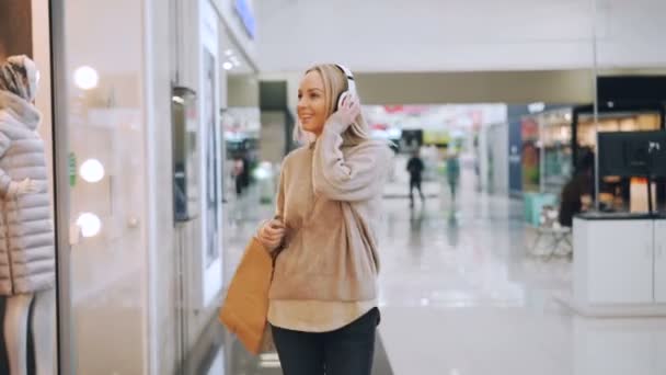 La guapa rubia se está divirtiendo en el centro comercial escuchando música a través de auriculares, bailando con bolsas de papel y mirando a su alrededor la nueva ropa de moda . — Vídeo de stock