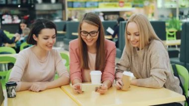 Neşeli genç bayanlar birlikte ekran gülümseyen seyir smartphone kullanıyorsanız ve alışveriş merkezi kağıt ile rahat kafe gülen oturma görünür torbalar. Gadget'lar ve eğlence kavramı.