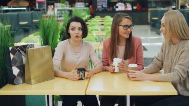 Şirin kız içecekler ve kağıt torbalar ile Alışveriş Merkezi kafede masada oturan ve paylaşım haber konuşuyor. Kadın trendy giyim ve aksesuarları üzerinde var.