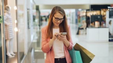 Cherful genç kadın smartphone ekran dokunaklı o zaman kağıt torba tutarak giyim yeni koleksiyon etrafa alışveriş merkezinde kullanıyor. Gençlik ve gadget'ları kavramı.