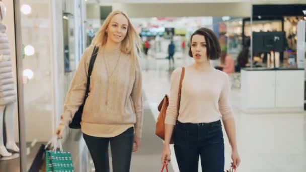 Las amigas jóvenes guapas están charlando y sonriendo disfrutando del día de compras en una gran tienda espaciosa mientras caminan en el pasillo. Las chicas llevan bolsas de papel.. — Vídeo de stock