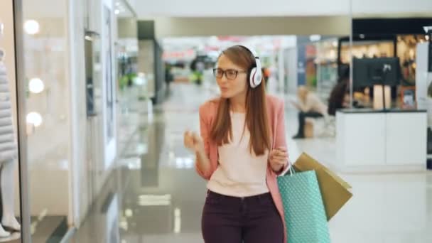 Mulher feliz está desfrutando de música em fones de ouvido, dançando e cantando em shopping center hall segurando sacos de papel. Menina está olhando para roupas em vitrines e se divertindo . — Vídeo de Stock