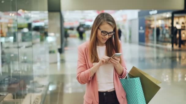 Roztomilá dívka s blond vlasy používá smartphone a usmíval se při chůzi v nákupní centrum s papírové sáčky. Internet, moderní technologie a koncepce životního stylu mládeže. — Stock video