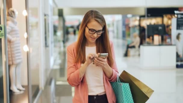 Cherful νεαρή γυναίκα χρησιμοποιώντας το smartphone στο εμπορικό κέντρο, αγγίζοντας την οθόνη και, στη συνέχεια, να βλέπουν γύρω από τη νέα συλλογή ενδυμασία κρατώντας χάρτινες σακούλες. Έννοια νέων και μικροεφαρμογές. — Αρχείο Βίντεο