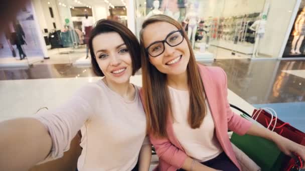 ショッピング モールとを持つ楽しい舌を示し、面白い顔を作るペーパー バッグと selfie を取って可愛い女の子友達の視点ショット。友情と技術の概念. — ストック動画