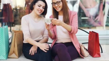 Duygusal kızlar sohbet ve alışveriş torbaları ile bankta oturmuş ve having fun gülüyor smartphone ekranında arıyoruz. Modern teknoloji ve iletişim kavramı.
