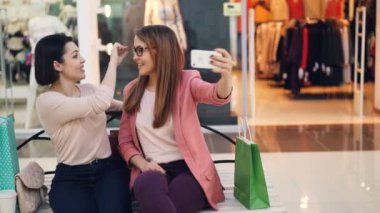 Neşeli bayan arkadaş selfie alışveriş merkezi bankta oturmuş ve komik surat gözlüklerle poz smartphone ile alıyor. Fotoğraf, mağazalar ve dostluk kavramı.