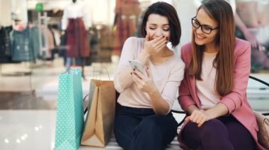 Neşeli genç kadınlar birlikte ekran gülümseyen seyir smartphone kullanıyorsanız ve kağıt ile alışveriş merkezi bankta gülen dinlenme görünür torbalar. Gadget'lar ve eğlence kavramı.