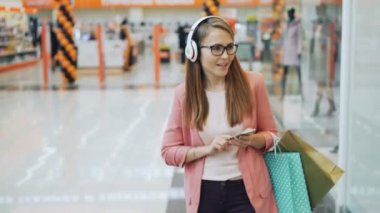 Güzel Bayan kulaklık müzik ve kağıt torbalar ile alışveriş merkezi yürüme smartphone kullanarak. Modern araçlar, millennials ve mutluluk kavramı.