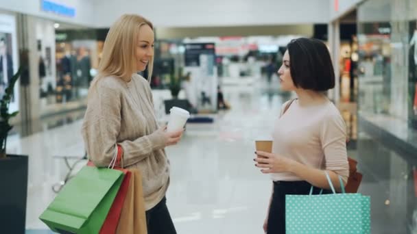 Hermosas mujeres jóvenes están charlando en el centro comercial con bebidas para llevar y bolsas de papel discutiendo compras y compartiendo noticias. Estilo de vida juvenil y concepto de tiendas . — Vídeos de Stock