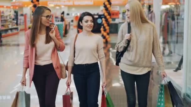 De jolies jeunes femmes parlent en marchant dans un centre commercial portant des sacs en papier avec des achats. Vente au détail, clients heureux et concept de style de vie des jeunes . — Video