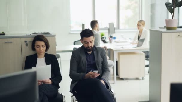 I giovani uomo e donna sono in attesa di un colloquio di lavoro in carica mentre il manager sta intervistando un altro candidato. Ragazza è in possesso di cv, ragazzo utilizza smartphone . — Video Stock