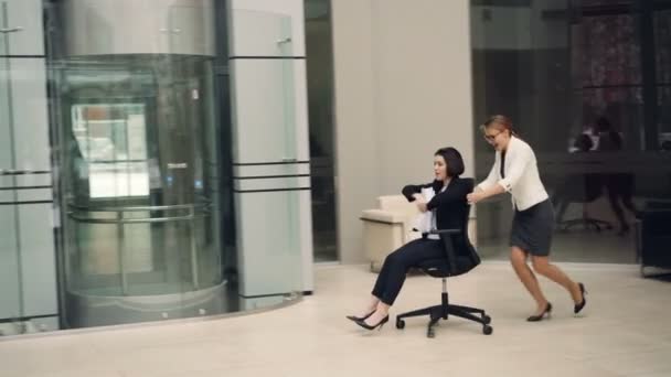 Speelse meisjes zakenvrouwen plezier in office lobby stoelen rijden en foute papieren genieten van vrijheid en einde aan werken. Jeugd, werkruimte en vreugde concept. — Stockvideo